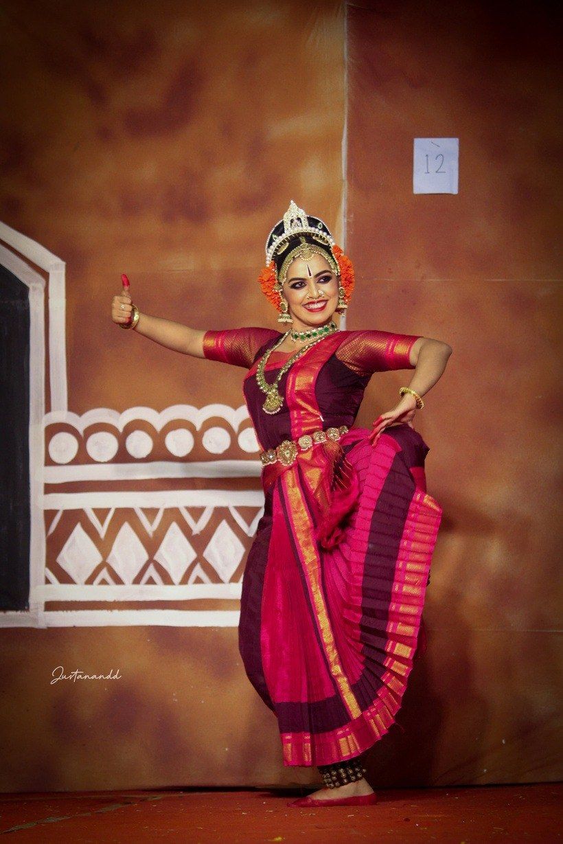 Medha Mayuri in magenta against a painted mural backdrop, arm extended.