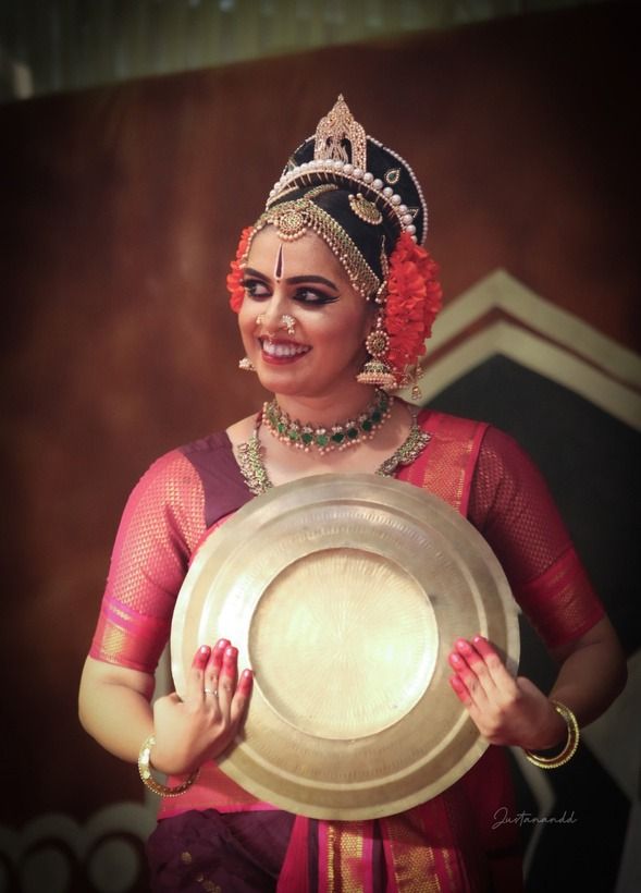 Medha Mayuri in a magenta and gold costume, holding a brass plate, smiling.